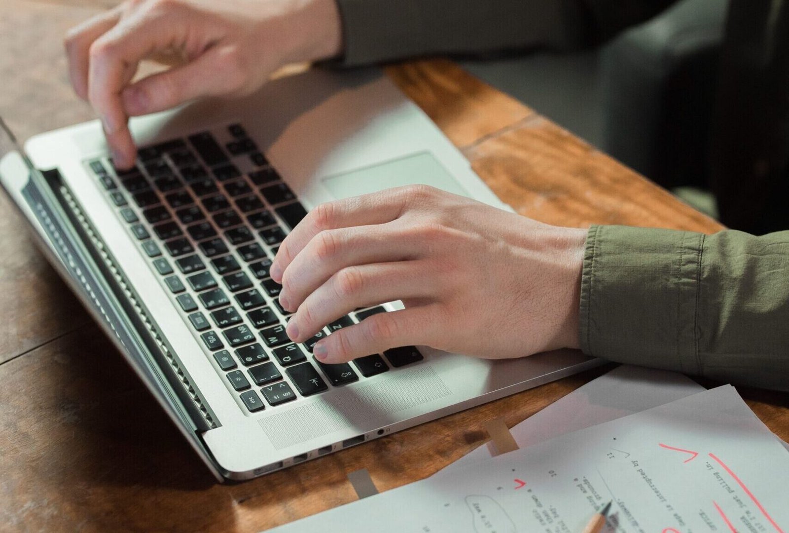 Focused person typing on laptop with notes and papers beside on wooden table.